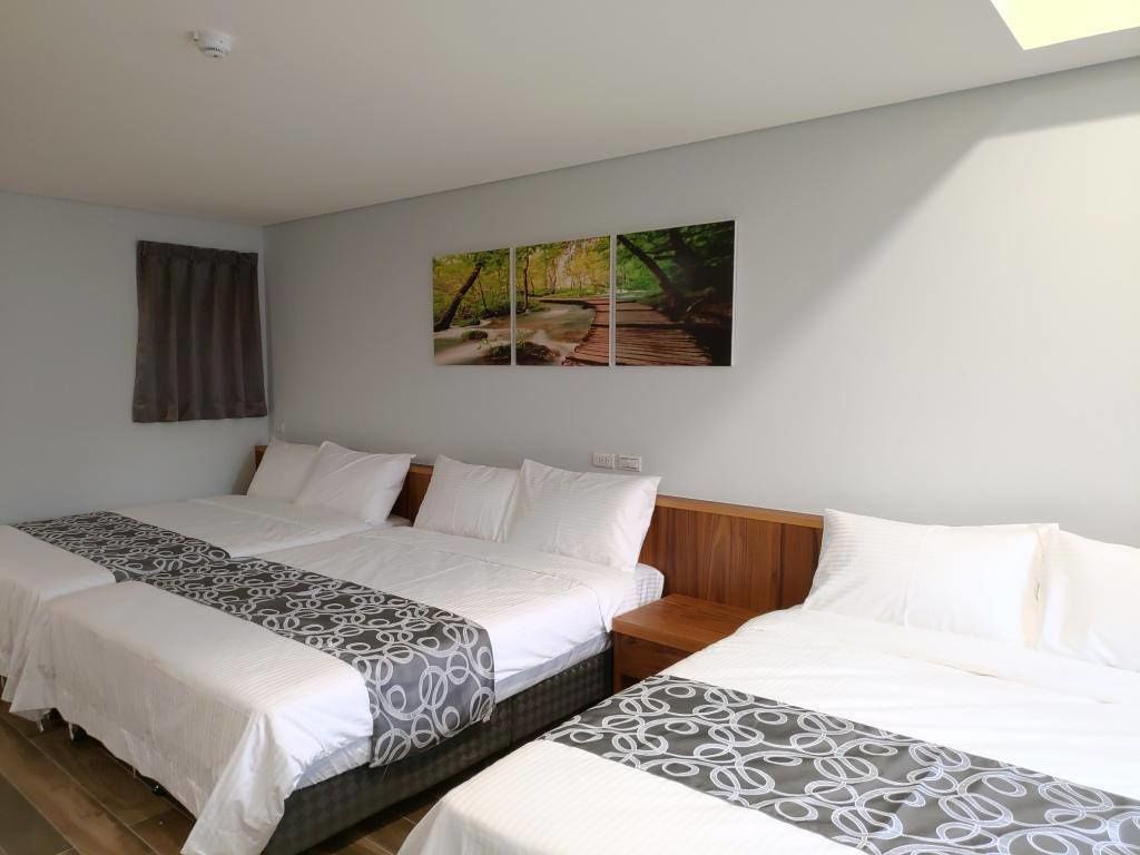 A hotel room with two beds, a wooden headboard, a painting of a forest path, and a window with curtains.