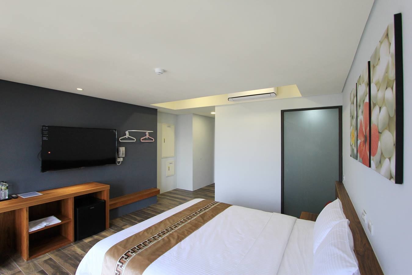 A modern hotel room with a large bed, a desk with a flat-screen TV, a wardrobe, and a door leading to another room.