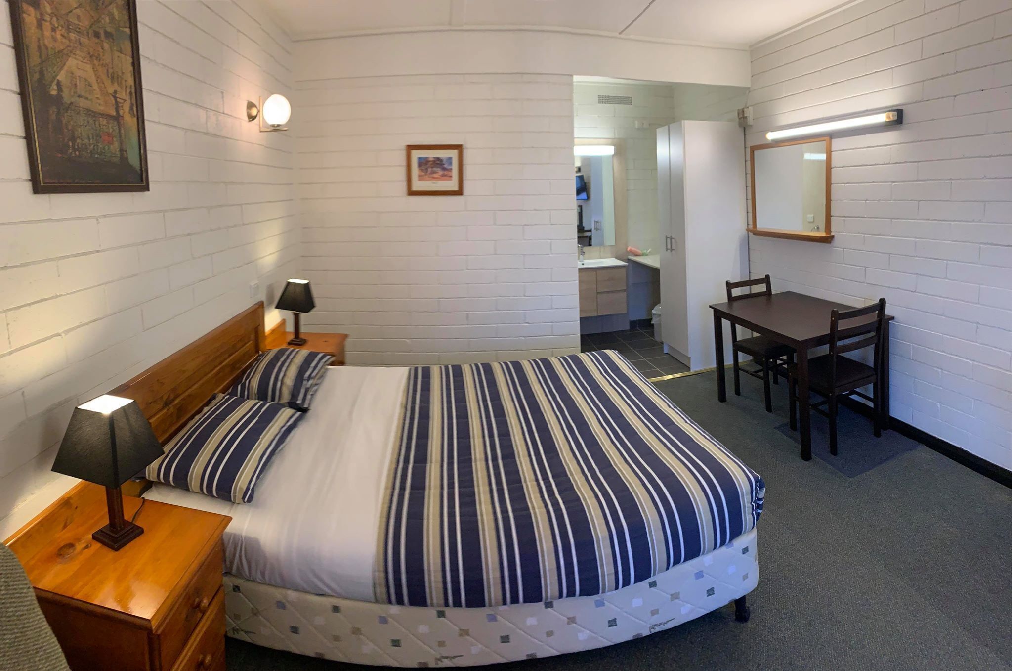 Quarto com cama, mesa de cabeceira, luminária e escrivaninha com cadeiras.