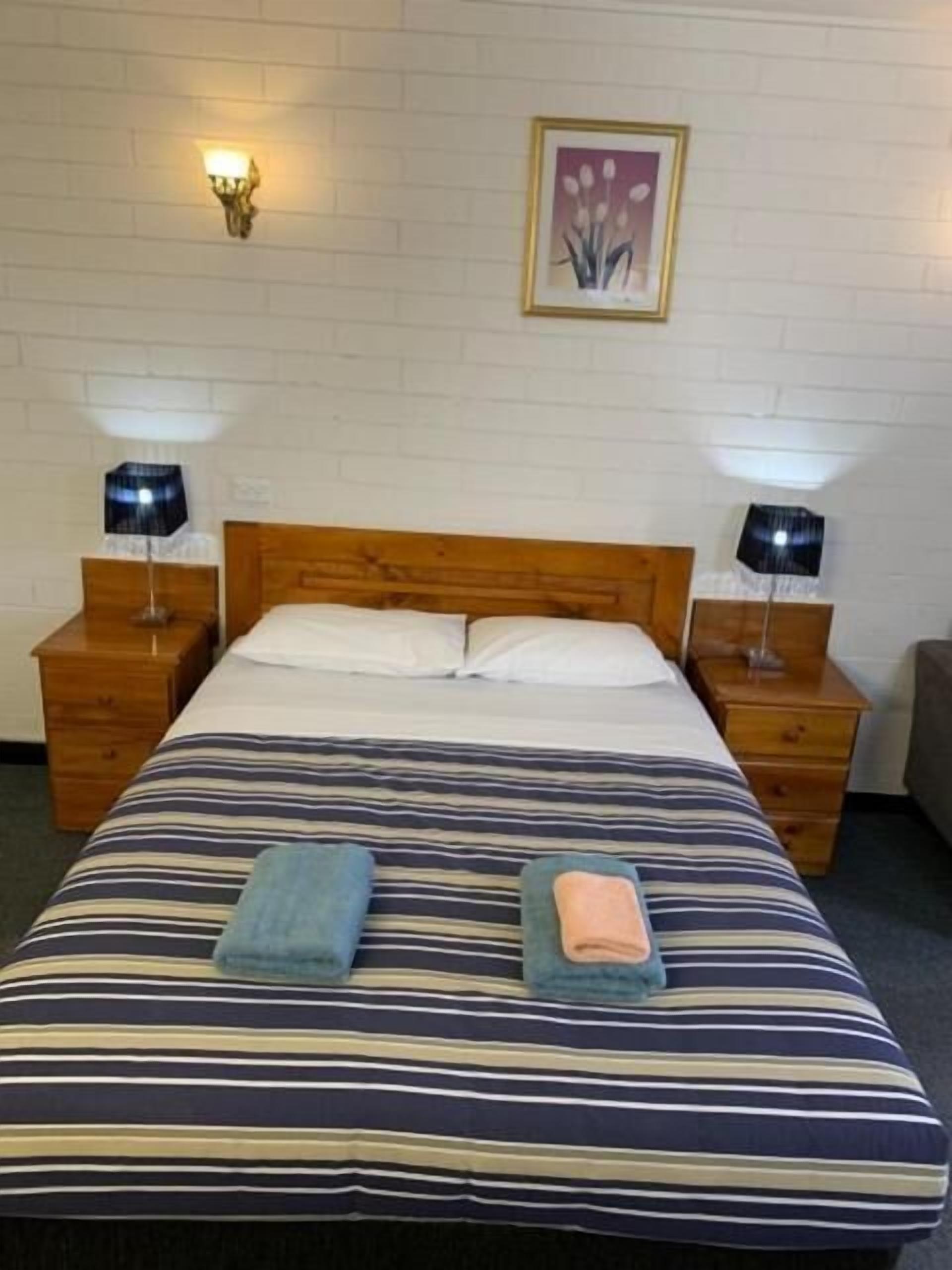 Een hotelkamer met een eenpersoonsbed, twee nachtkastjes met lampen, een lijst aan de muur en een klein houten nachtkastje.