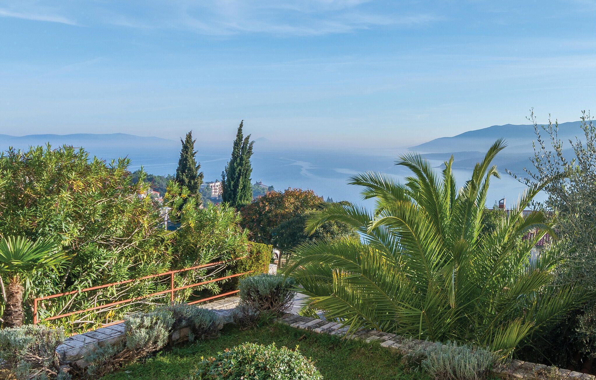 Un jardín arbolado con un sendero de piedra, palmeras y vistas al mar y montañas distantes.