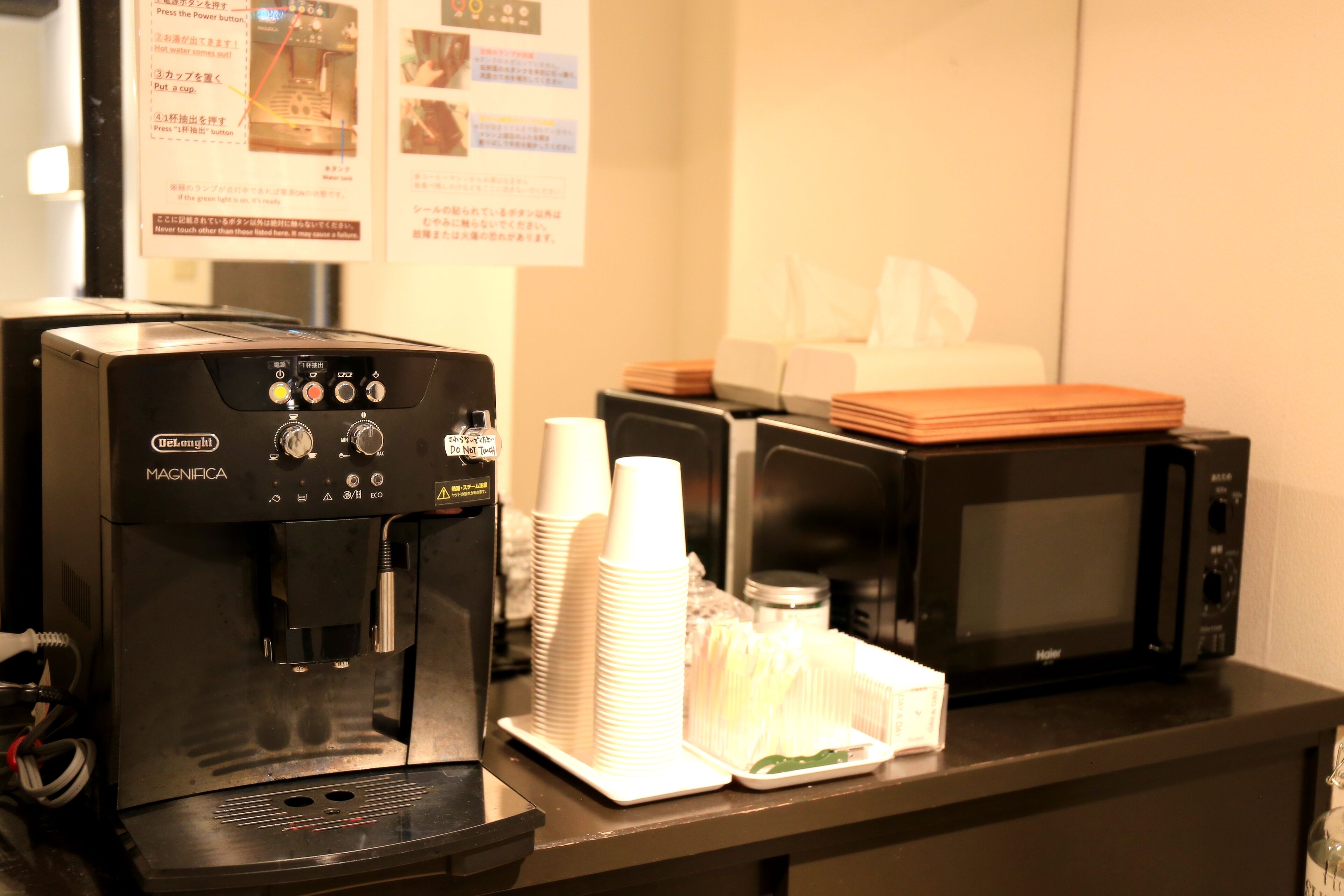 Uma máquina de café, pilhas de copos descartáveis, um micro-ondas e uma caixa de lenços em uma bancada.
