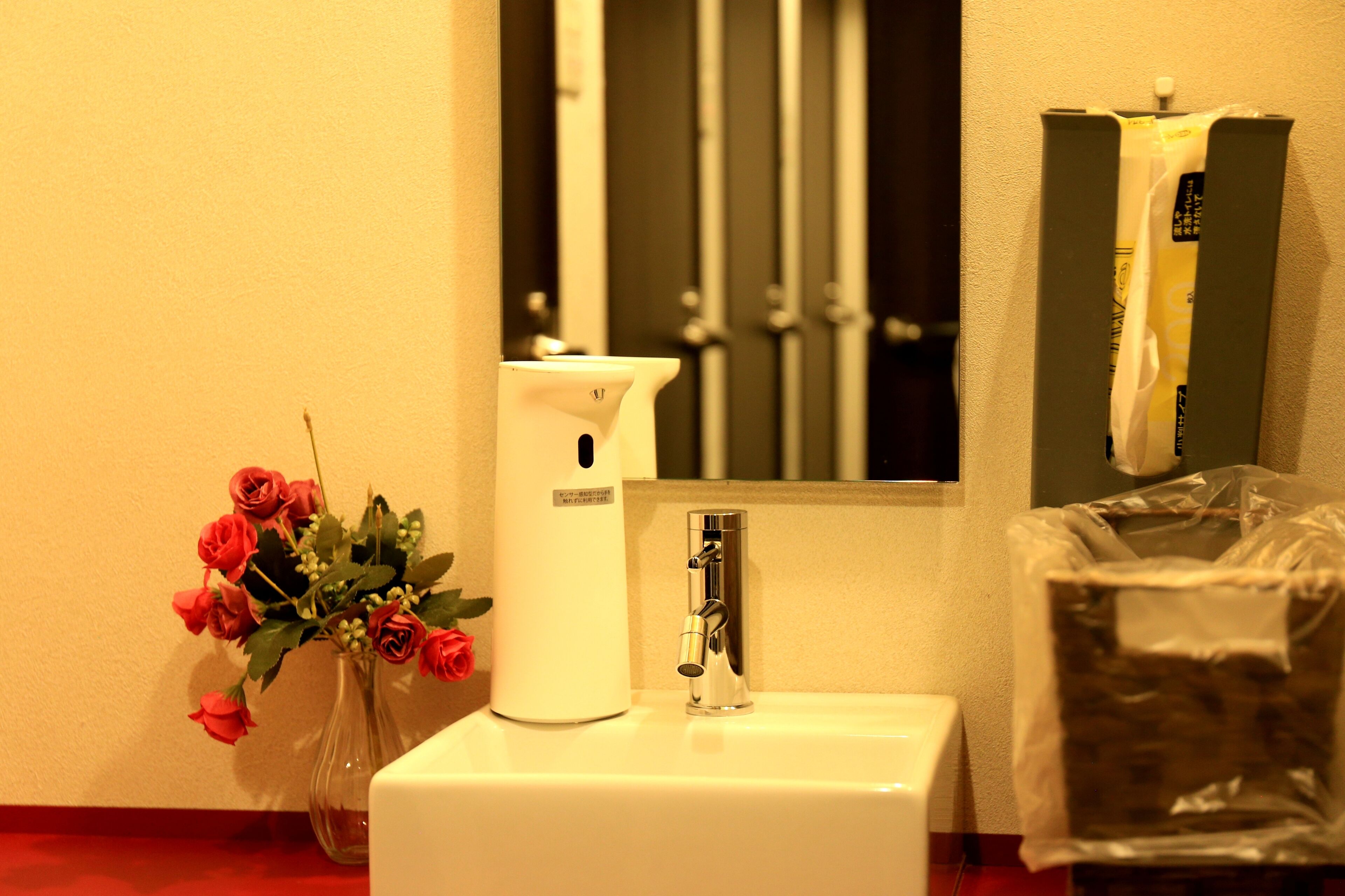 Un baño con lavamanos blanco, grifo, un florero con rosas rojas y un secador de manos.