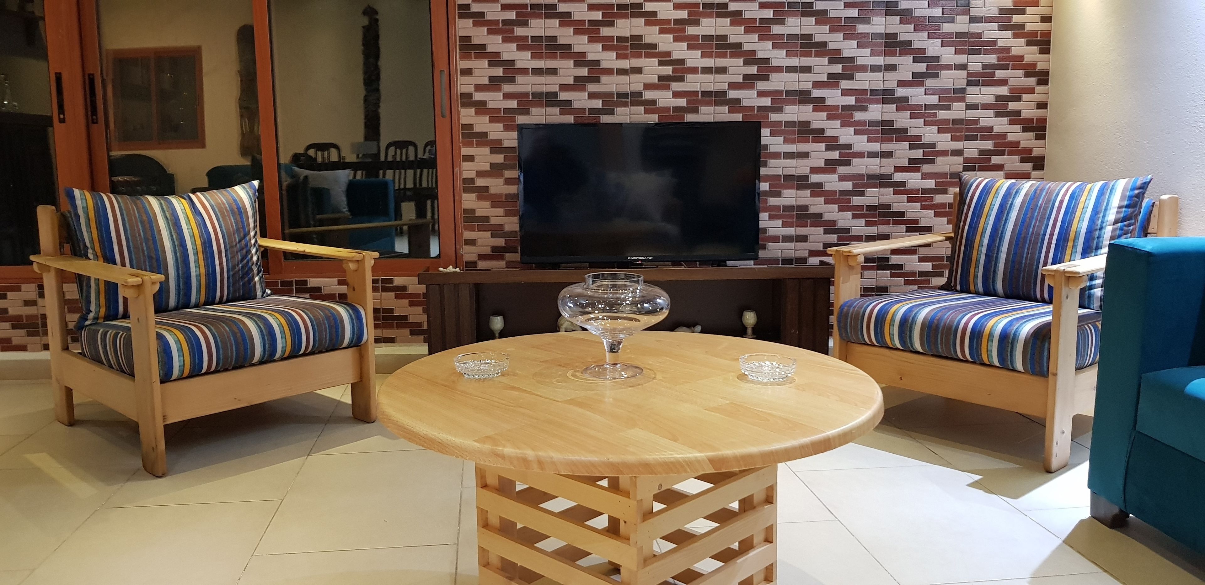 A living room with two striped armchairs, a round wooden table with two glass bowls, and a flat-screen TV against a brick-patterned wall.