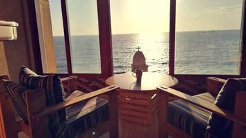 Une pièce avec une table ronde en bois et deux chaises à coussins rayés, une fenêtre donnant sur la mer et un navire, et une petite table avec une tasse.