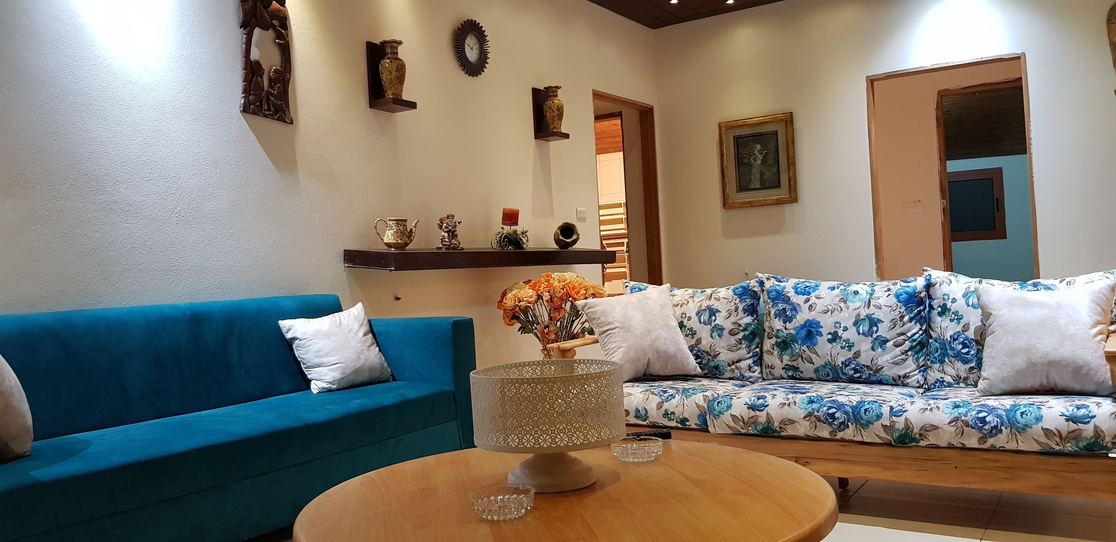 Un salón con un sofá azul, un sofá floral, una mesa de centro y objetos decorativos en estanterías.