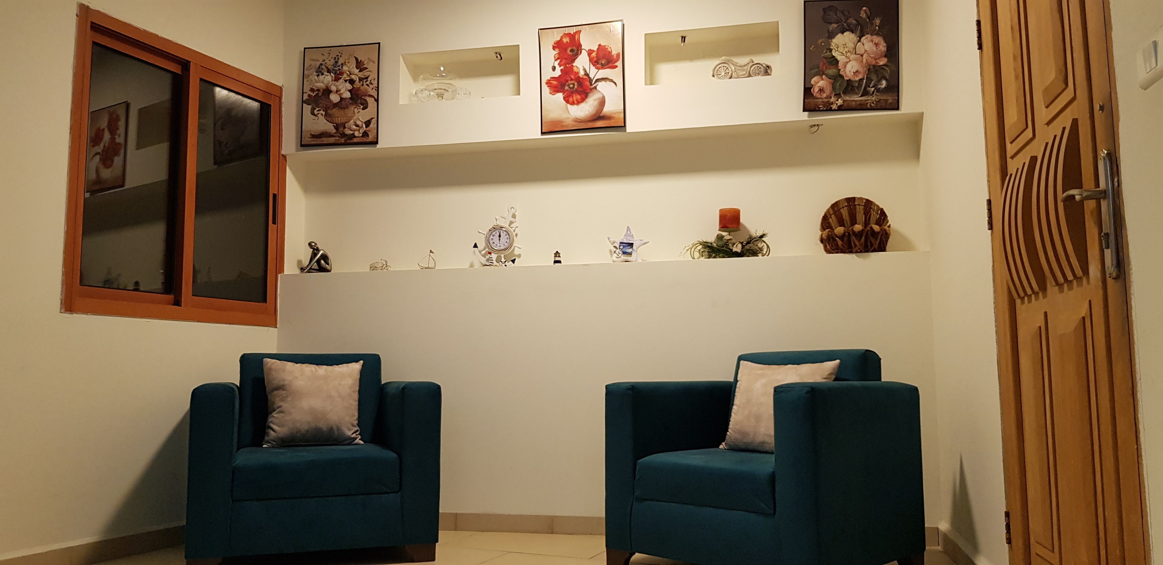 A room with two teal armchairs, a wooden door, and a white wall with shelves holding various decorative items.
