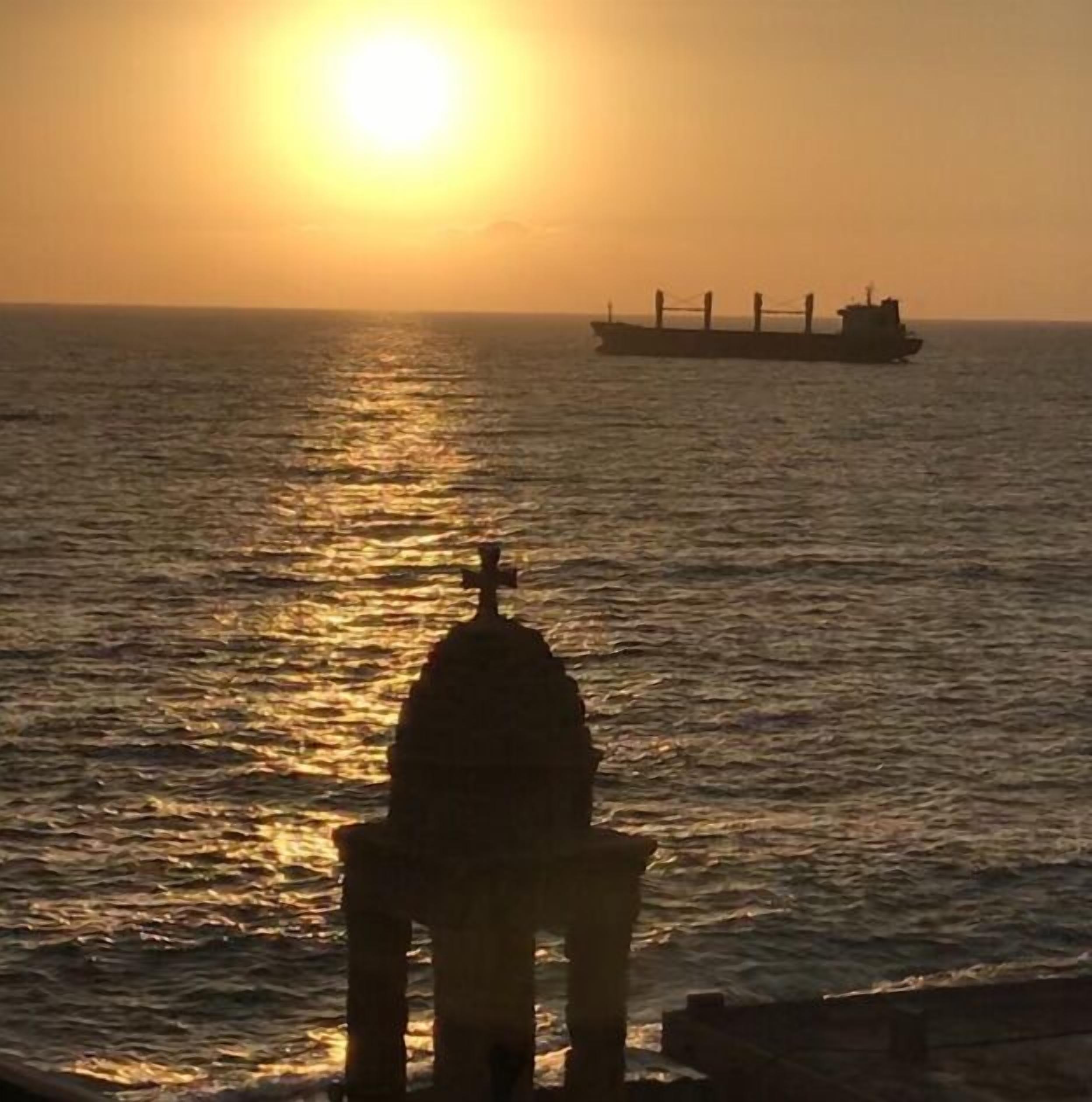 Een zonsondergang boven de oceaan, met een schip in de verte en een gebouw met een koepel op de voorgrond.