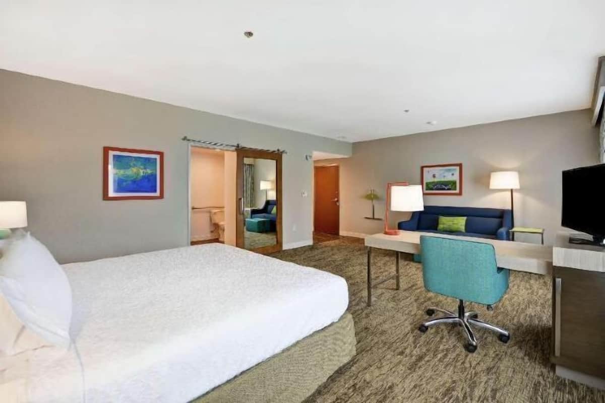 A hotel room with a bed, a desk, a chair, a TV, and a door leading to another room.