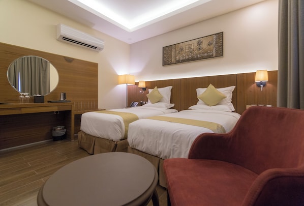 Une chambre d’hôtel avec deux lits, une table ronde, un fauteuil rouge, une tête de lit en bois, un climatiseur fixé au mur et une œuvre d’art encadrée au-dessus des lits.
