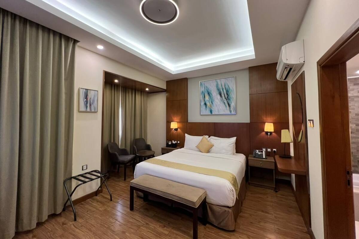 Habitación de hotel con una cama grande, mesitas de noche, un escritorio, una silla, un banco, una lámpara de techo y un equipo de aire acondicionado.