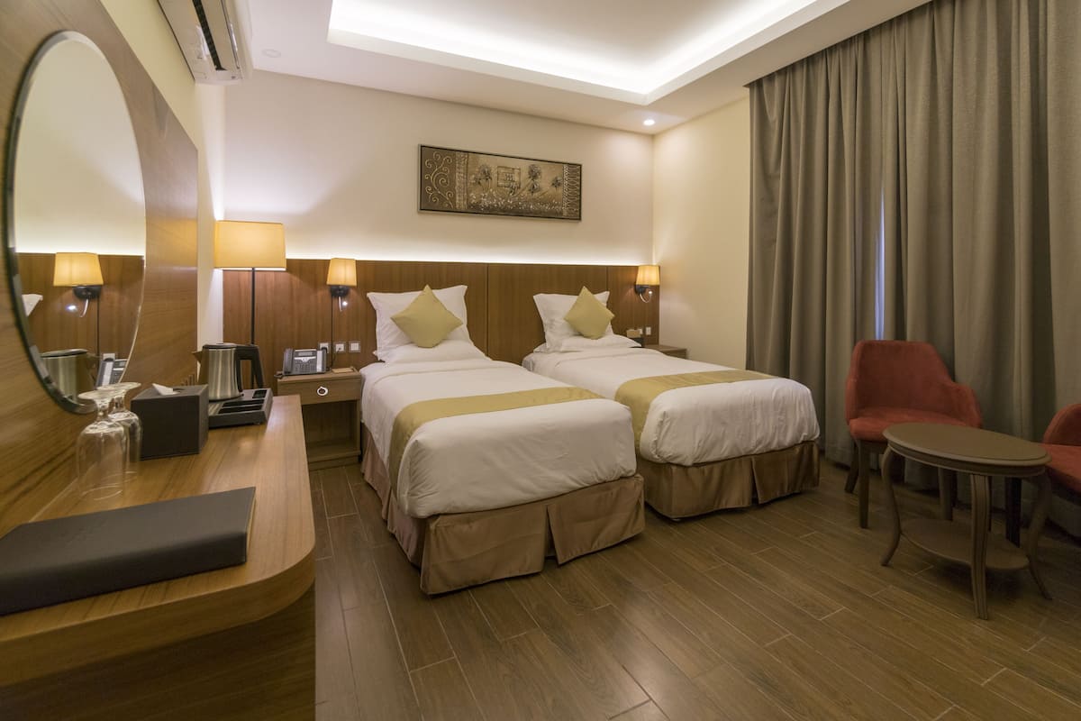 Habitación de hotel con dos camas, un escritorio, una silla, una mesita, un espejo y un cuadro en la pared.