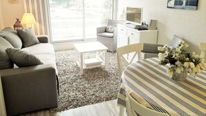 A living room with a sofa, a small table, a TV, and a dining area with a striped tablecloth. - Beautiful 2 rooms Moulleau center with parking (ARCACHON)