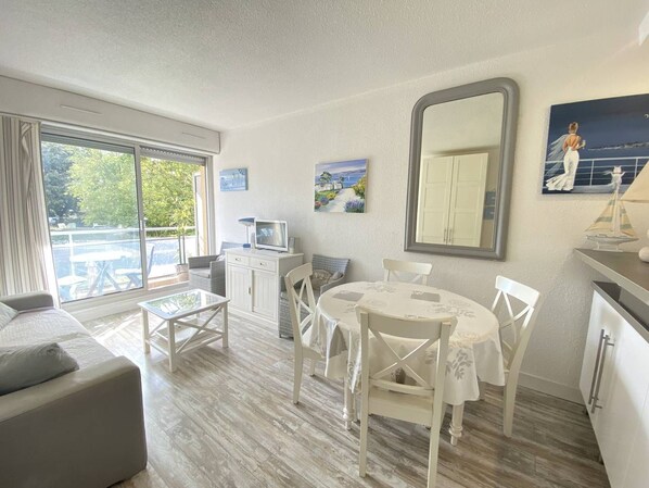 Een kamer met een balkon, een eettafel met stoelen, een bank, een televisie en een grote spiegel.