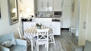 A compact kitchen with white cabinets, a microwave, and a small dining area with striped tablecloths and white chairs. - Beautiful 2 rooms Moulleau center with parking (ARCACHON)