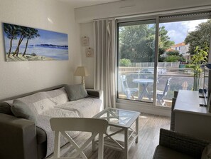 Een kamer met een bank, een tafel met glazen blad en uitzicht op een parkeerplaats door een groot raam.