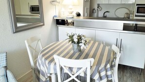A compact kitchen with white cabinets, a microwave, a sink, and a dining area with a striped tablecloth and white chairs. - Beautiful 2 rooms Moulleau center with parking (ARCACHON)