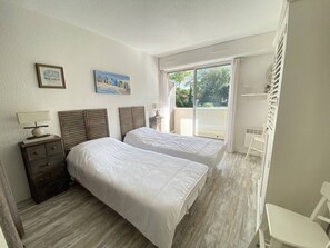 A bedroom with two beds, a nightstand, a lamp, a painting, and a view of the outdoors. - Beautiful 2 rooms Moulleau center with parking (ARCACHON)