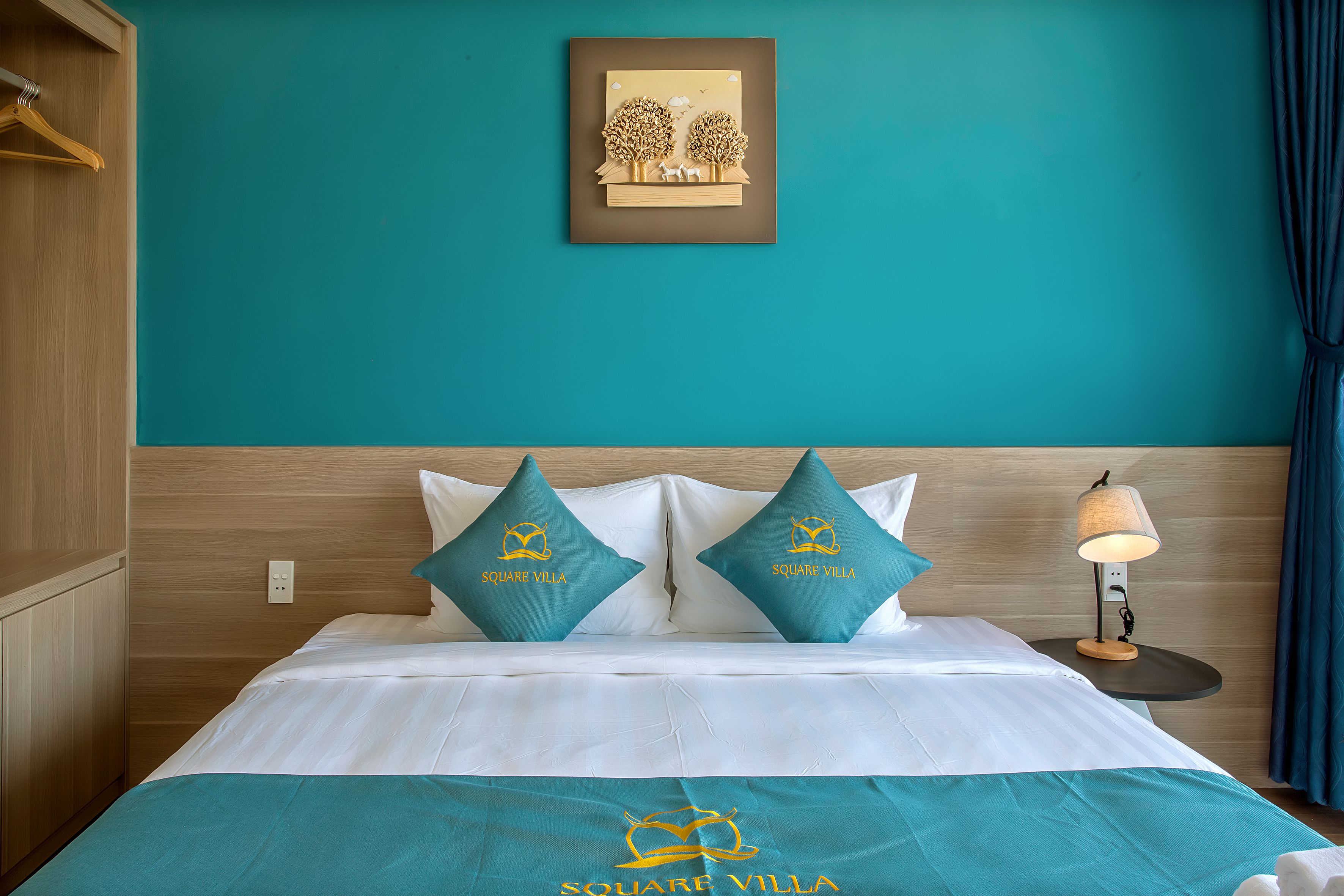 A hotel room with teal walls, a bed with white bedding, two teal pillows, a framed picture on the wall, and a nightstand with a lamp.