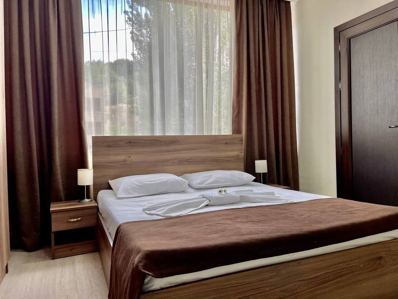 A neatly made bed with a brown headboard, two bedside tables with lamps, and a window with sheer curtains.