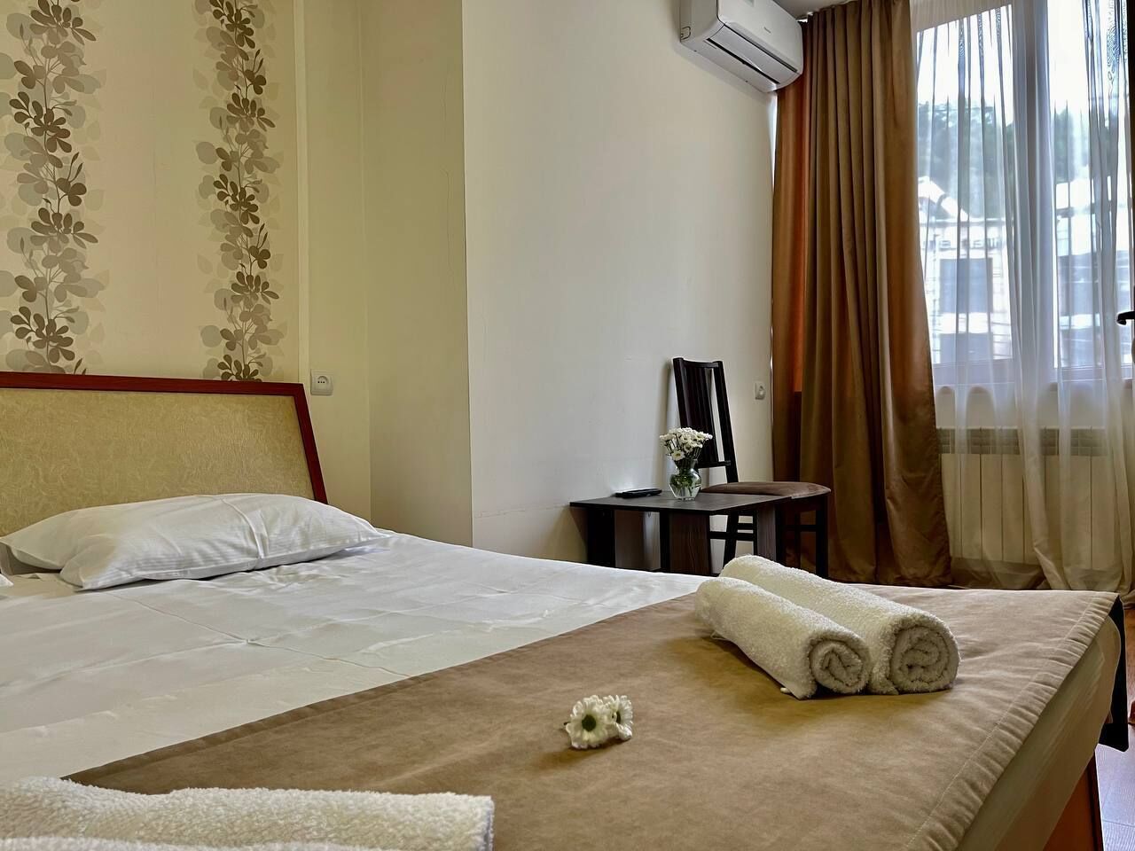 Une chambre d’hôtel avec un lit, une petite table sur laquelle est posé un vase de fleurs, et une vue sur l’extérieur à travers des rideaux légers.