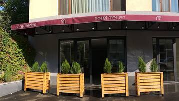 Een hotelentree met een bordeauxrode luifel, houten plantenbakken met groen en een heldere lucht.