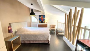 A bedroom with a large bed, a nightstand, a chair, a wooden chest, and a window with bamboo railings.