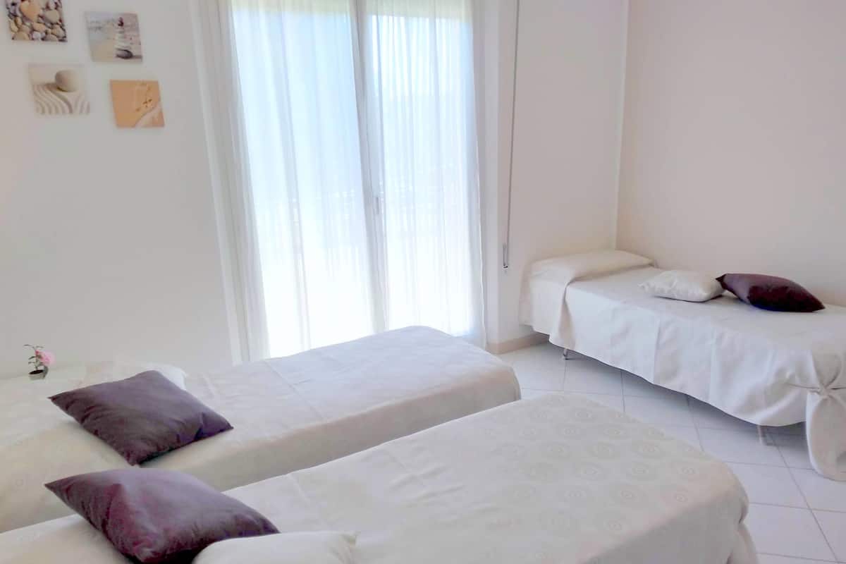 A room with two beds, each with a white mattress and a purple pillow. There is a window with sheer curtains, and the walls are plain white.