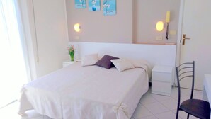 Uma cama bem arrumada com colcha e almofadas brancas, uma pequena mesa de cabeceira com um vaso de flores e uma obra de arte na parede acima da cama.