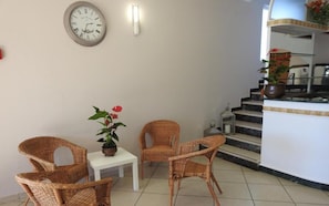 A room with wicker chairs, a small table, a clock, and a staircase leading to a balcony with plants.