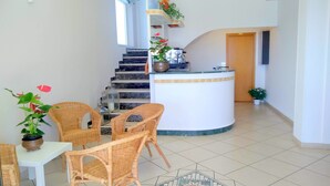 Um saguão de hotel com balcão de recepção, cadeiras de vime, piso ladrilhado e escada.