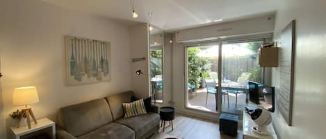 Ein modernes Wohnzimmer mit einer Couch, einem Holztisch und einem Essbereich. Eine Glastür öffnet sich zu einer Terrasse mit Tisch und Stühlen.