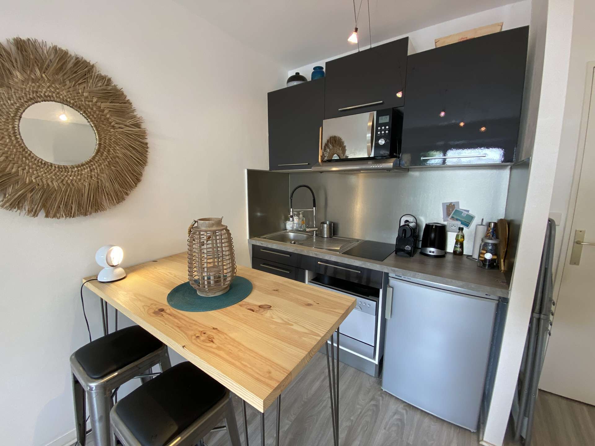 A compact kitchen with a wooden table, two bar stools, a microwave, a coffee maker, and a kettle.