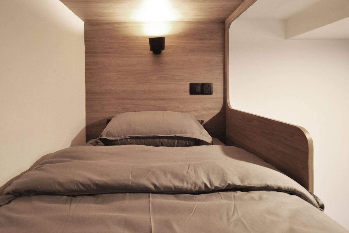 A compact bedroom with a wooden headboard, a single bed with beige bedding, and a wall-mounted light fixture.