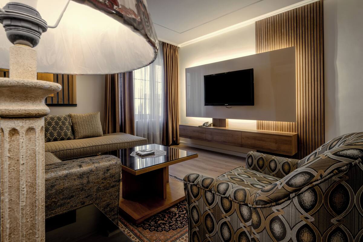 A modern living room with a sofa, armchair, and a coffee table. A flat-screen TV is mounted on the wall, and there is a decorative lamp.
