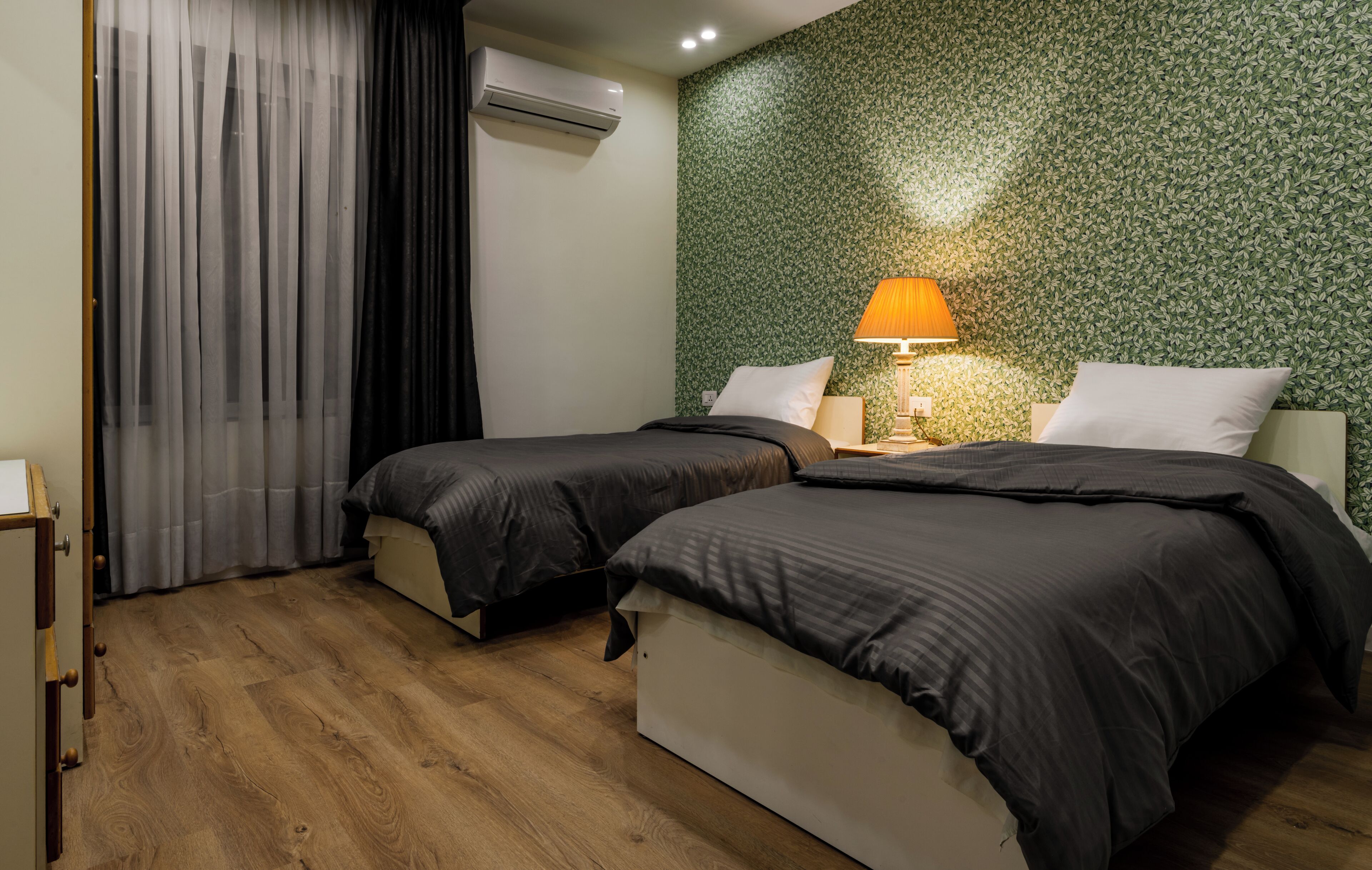 A hotel room with two beds, a wooden floor, a green leafy wallpaper, and a lamp on the nightstand.