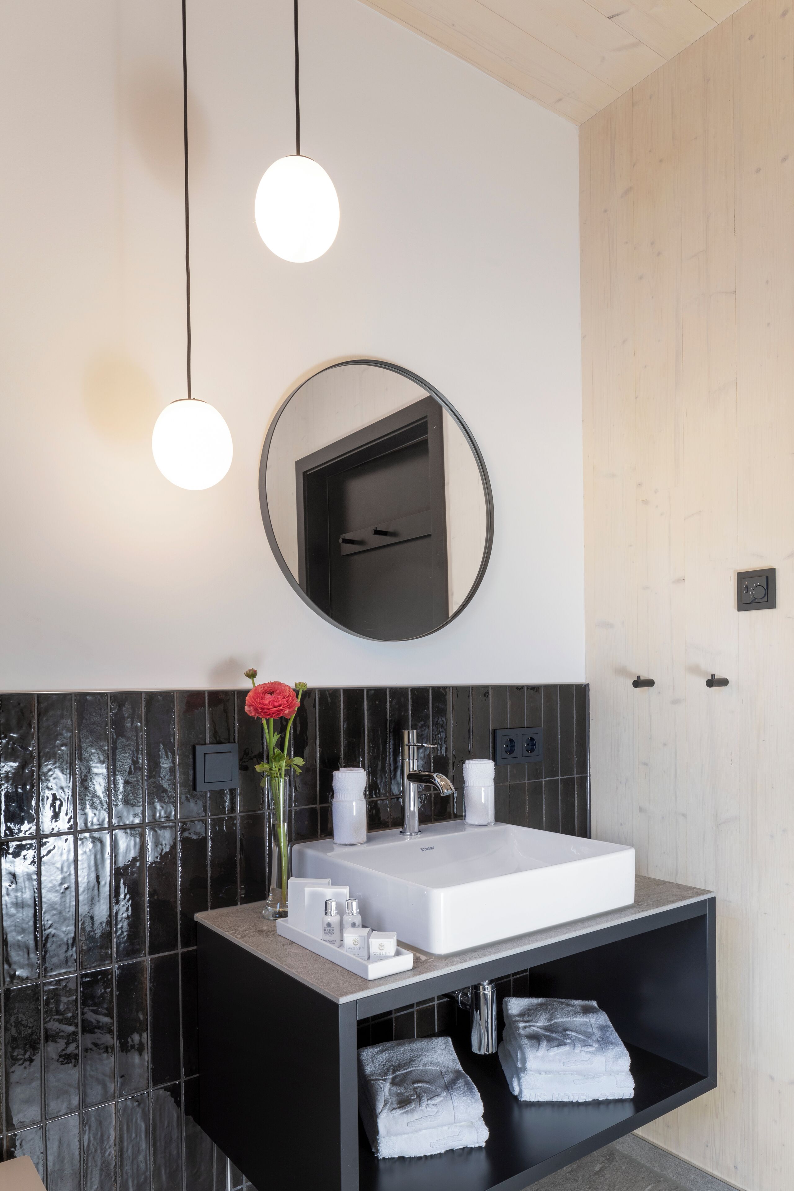 Un baño moderno con un lavamanos grande rectangular, un espejo redondo encima y un mueble negro debajo. Hay dos toallas sobre el mueble y un jarrón con una flor en el lavamanos.