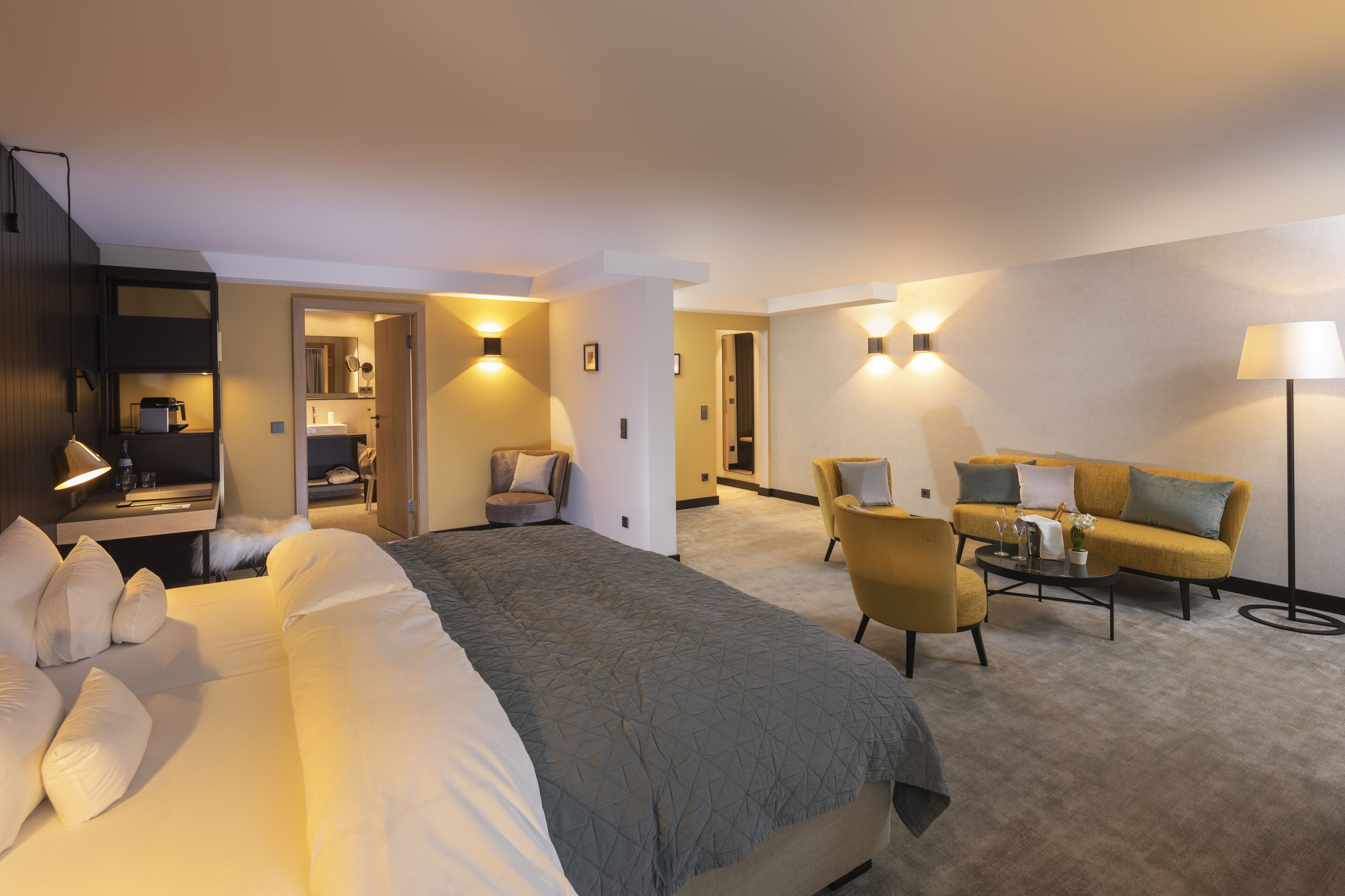 Ein Hotelzimmer mit einem großen Bett, einer Sitzecke, einem Couchtisch und einem Schreibtisch mit einer Lampe. Wandscheinwerfer sorgen für Beleuchtung.