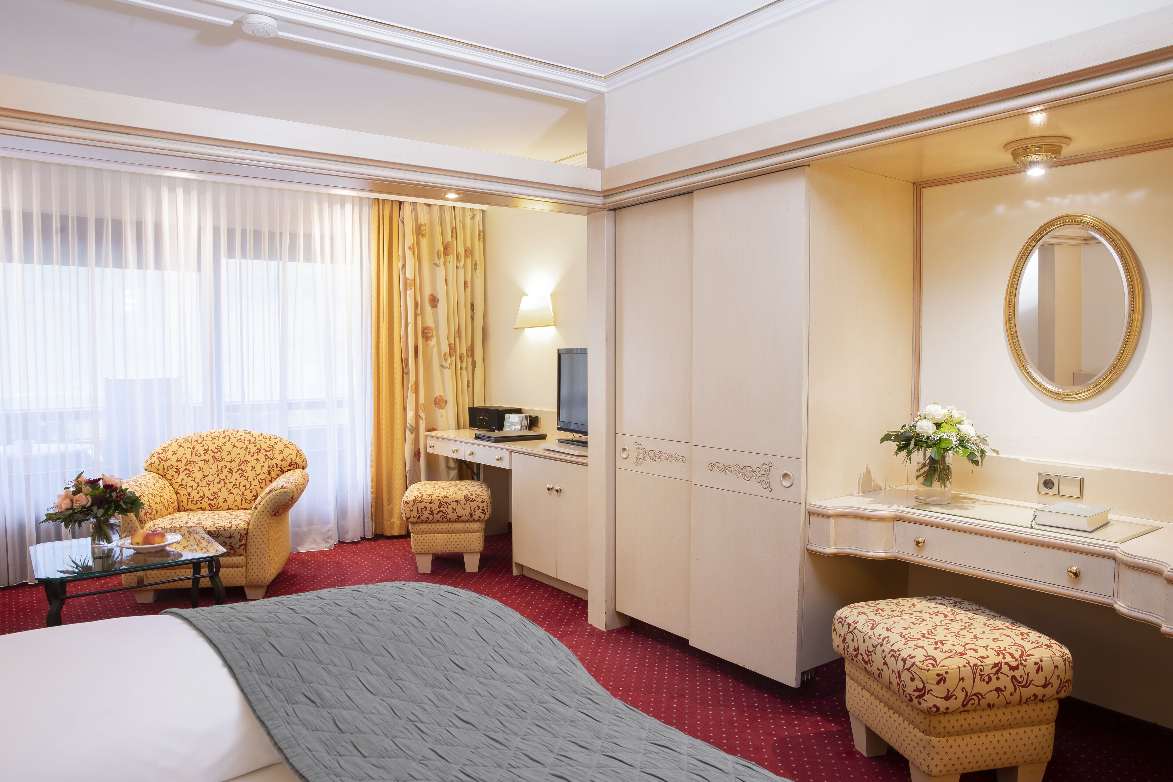 A hotel room with a bed, a desk with a computer, a chair, a small table with a vase, and a mirror.