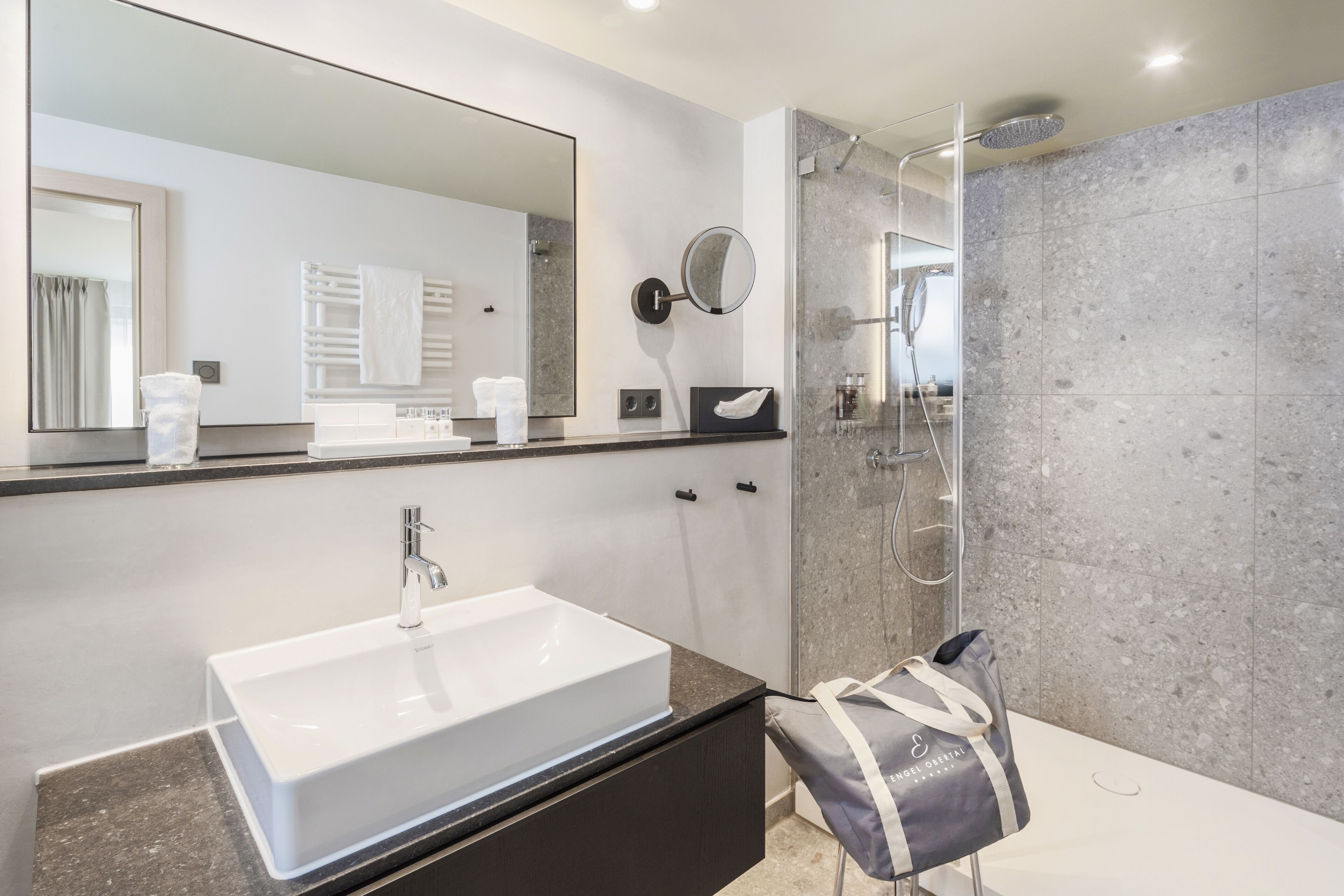 A modern bathroom with a large mirror, a white sink, a shower, and a towel rack.