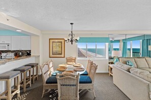 A modern kitchen with white cabinets, a dining area with wicker chairs and a glass table, and a living room with a beige sofa and a view of the ocean.