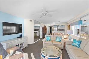 A living room with a flat-screen TV, a ceiling fan, a sofa with tropical pillows, and a wicker coffee table.