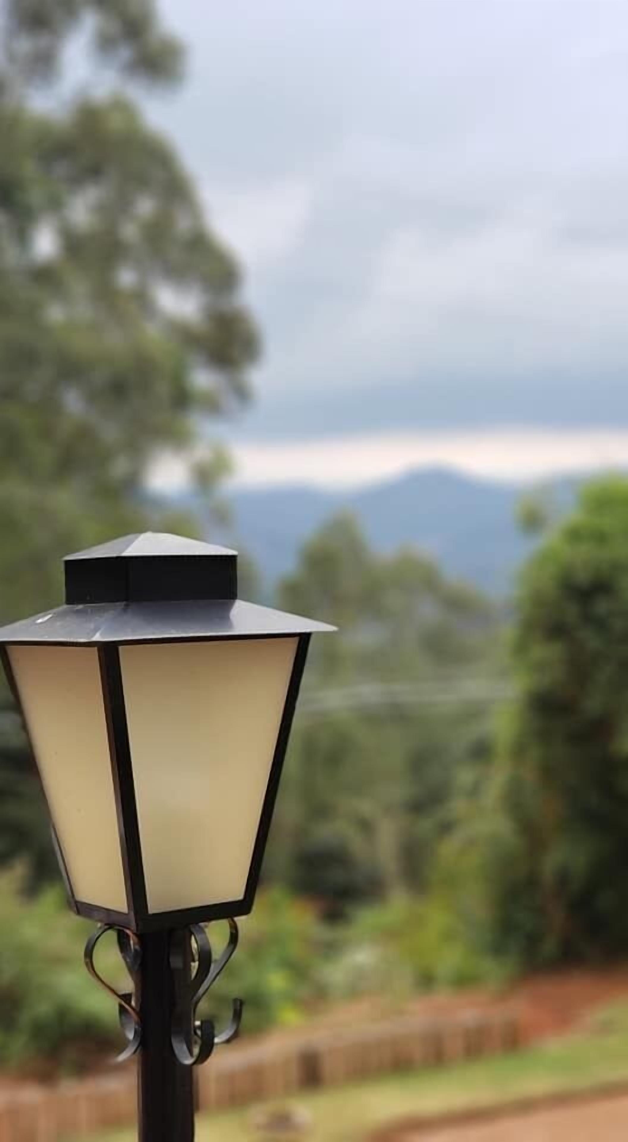 Una farola con base hexagonal y parte superior negra, frente a un fondo de vegetación y montañas lejanas.