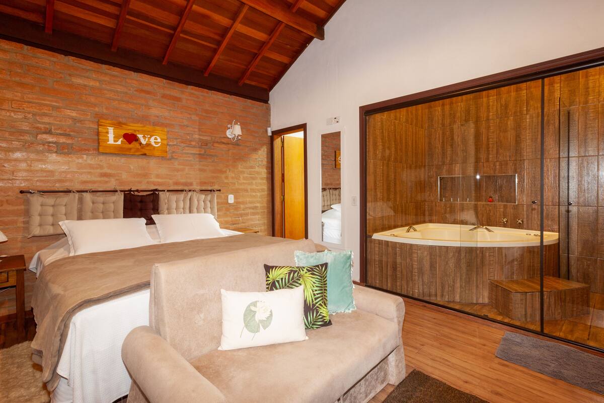 Quarto com uma cama grande, cabeceira de madeira, placa na parede com a inscrição 'Love' e casa de banho com amplas janelas e banheira.
