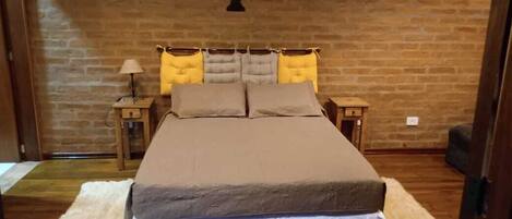 A bedroom with a brick wall, a bed with a white and brown bedspread, two nightstands with lamps, and a floral wall decoration.