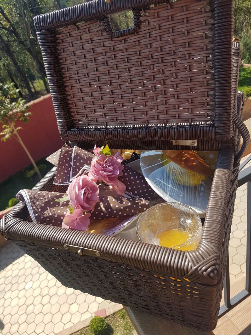 A wicker basket with a lid, containing a polka-dotted napkin, pink flowers, and a glass bowl with a yellow substance.