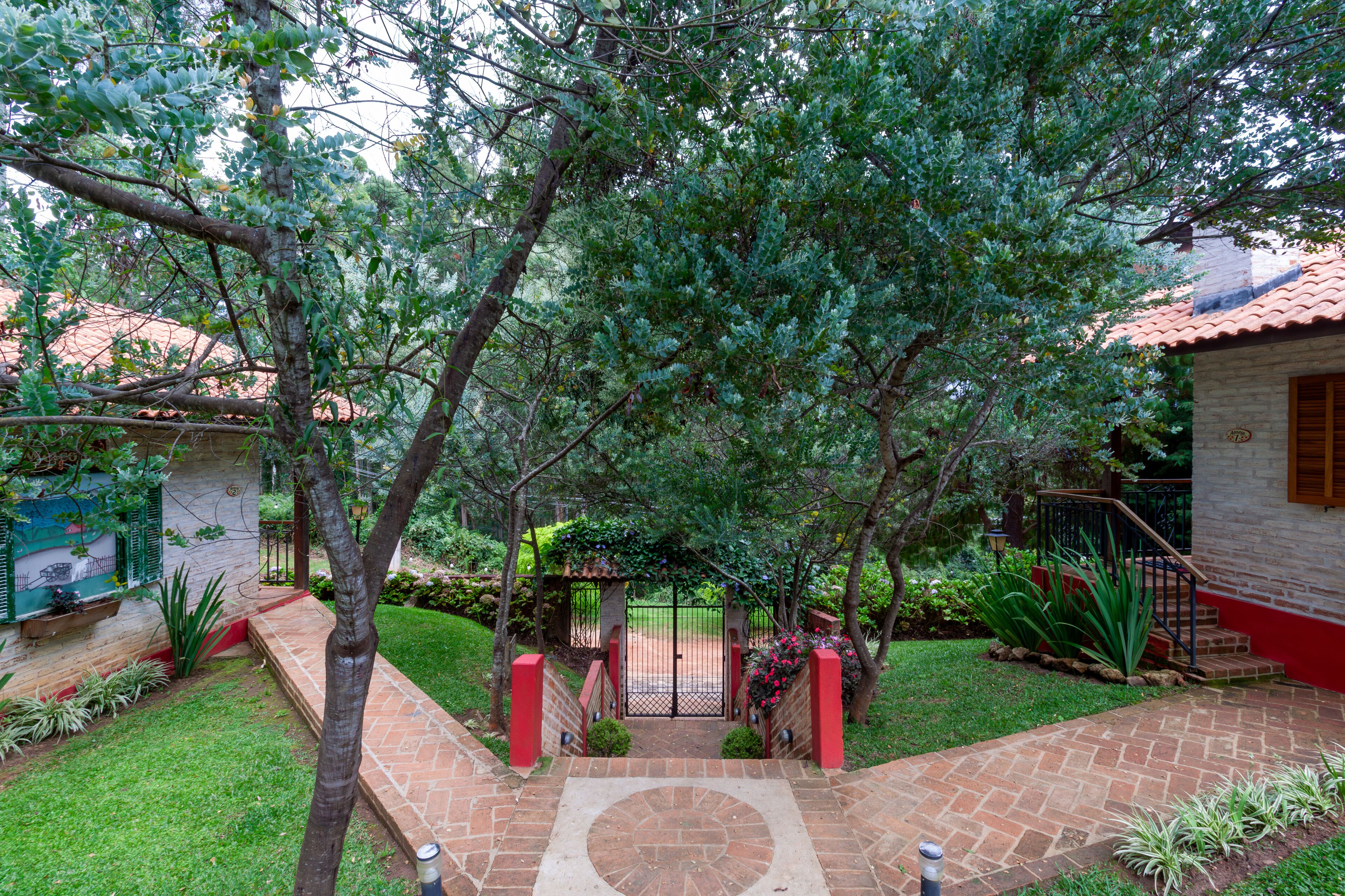 A courtyard with a brick pathway leading to a gated entrance, surrounded by lush greenery and a small building with a red roof.