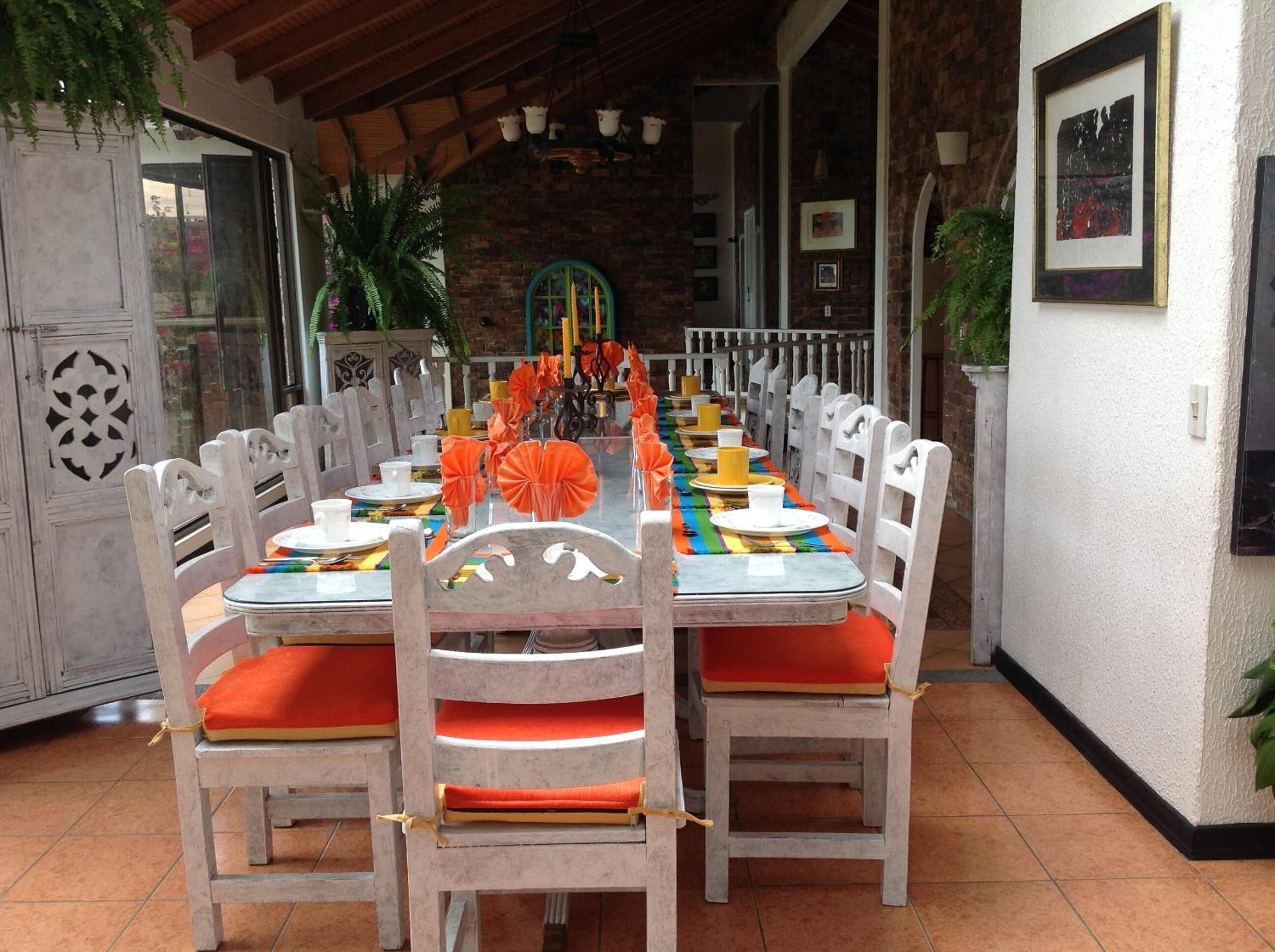 A dining area with a long table set for multiple people, surrounded by white chairs with orange cushions.