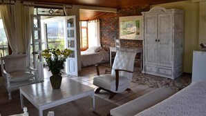 Un salón acogedor con un sofá blanco, un sillón blanco, una mesa blanca y un aparador blanco. Se aprecia una ventana con cortinas, un florero con flores y un cuadro en la pared.