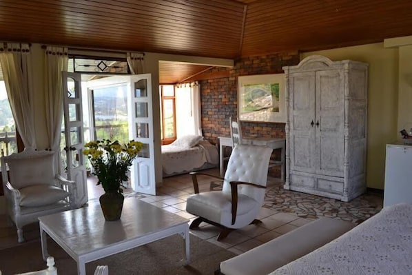Un salón acogedor con un sofá blanco, un sillón blanco, una mesa blanca y un aparador blanco. Se aprecia una ventana con cortinas, un florero con flores y un cuadro en la pared.